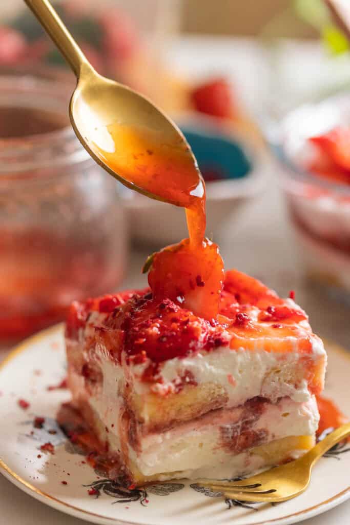 A slice of a Strawberry shortcake tiramisu on a cake plate with strawberry compote drizzling over the top.