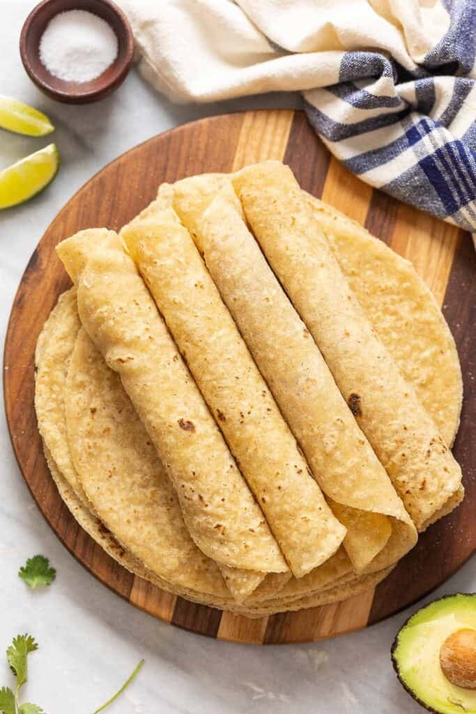 A wooden serving board with homemade gluten free flour tortillas - four tortillas on top are rolled up tightly like taquitos. 