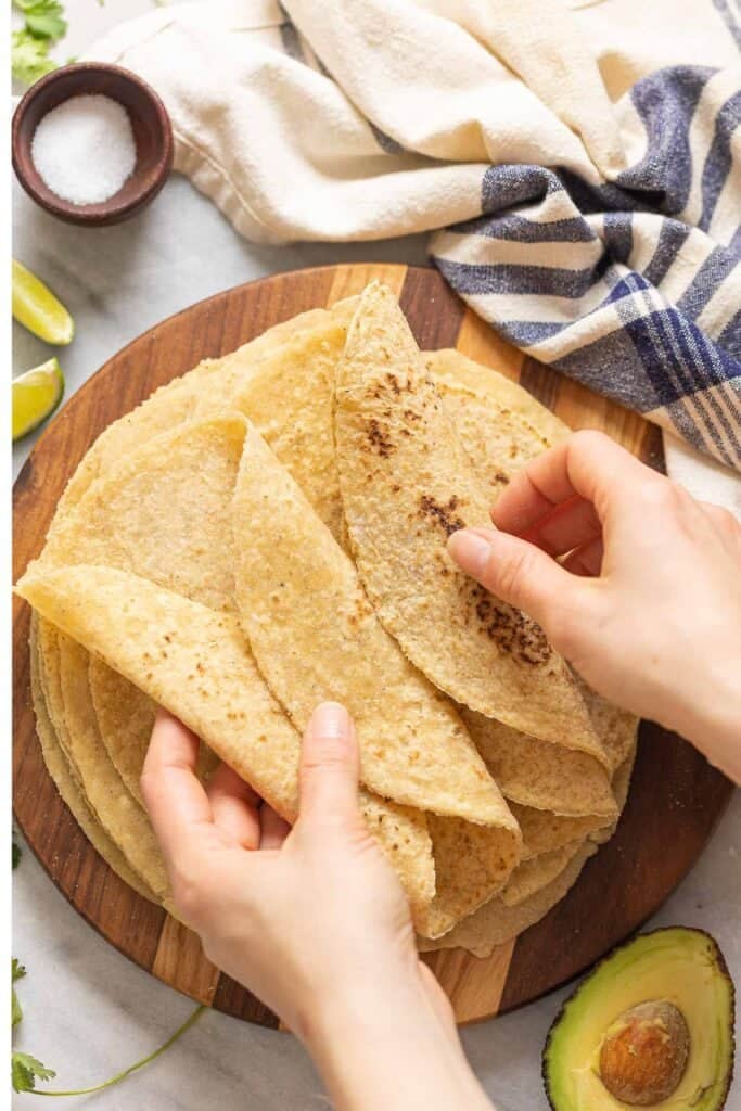 A wooden serving board with homemade gluten free flour tortillas. A pair of hands folding the top tortillas.