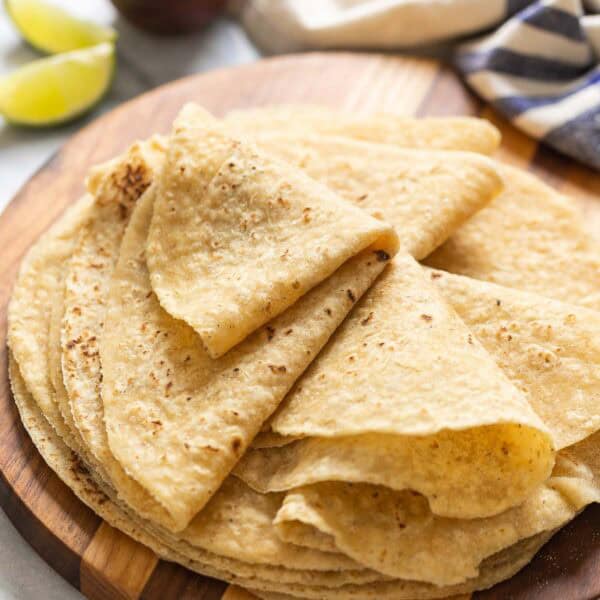 A wooden serving board with homemade gluten free flour tortillas on top. The top tortillas are folded up in wedges. Lime wedges and salt bowl in the background.