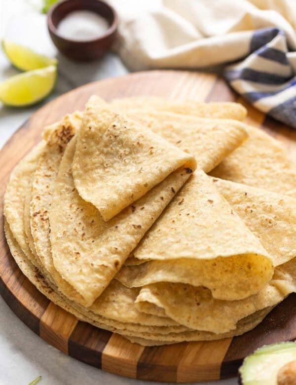 A wooden serving board with homemade gluten free flour tortillas on top. The top tortillas are folded up in wedges. Lime wedges and salt bowl in the background.