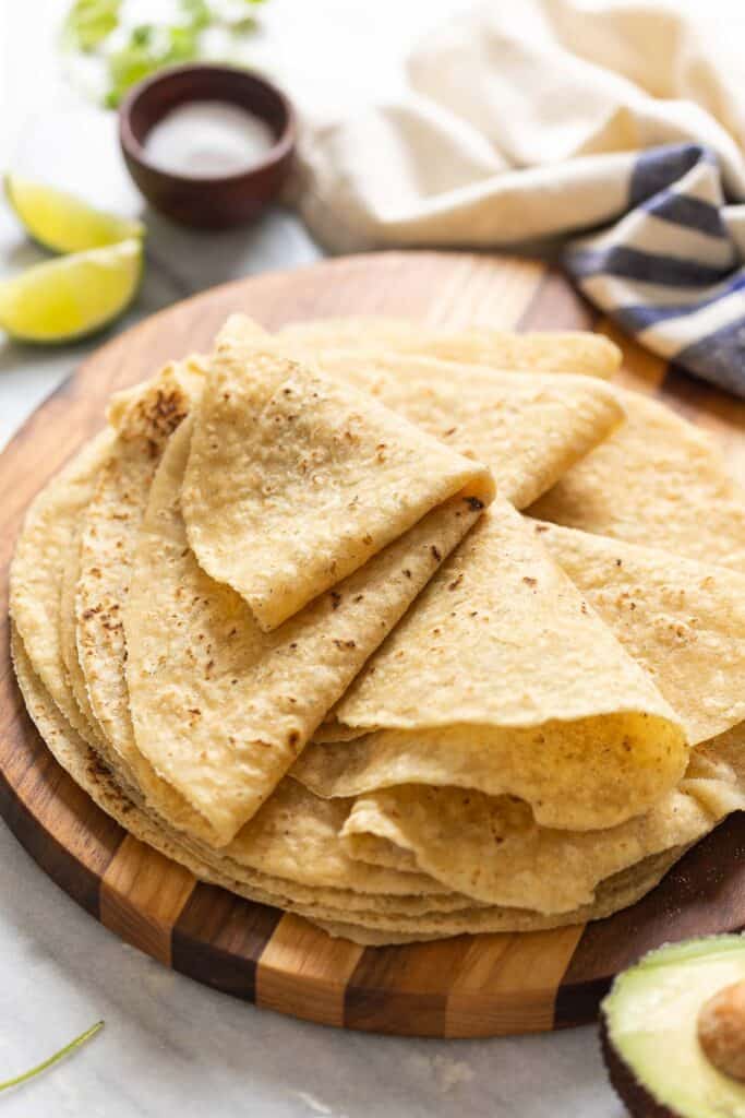 A wooden serving board with homemade gluten free flour tortillas on top. The top tortillas are folded up in wedges. Lime wedges and salt bowl in the background.