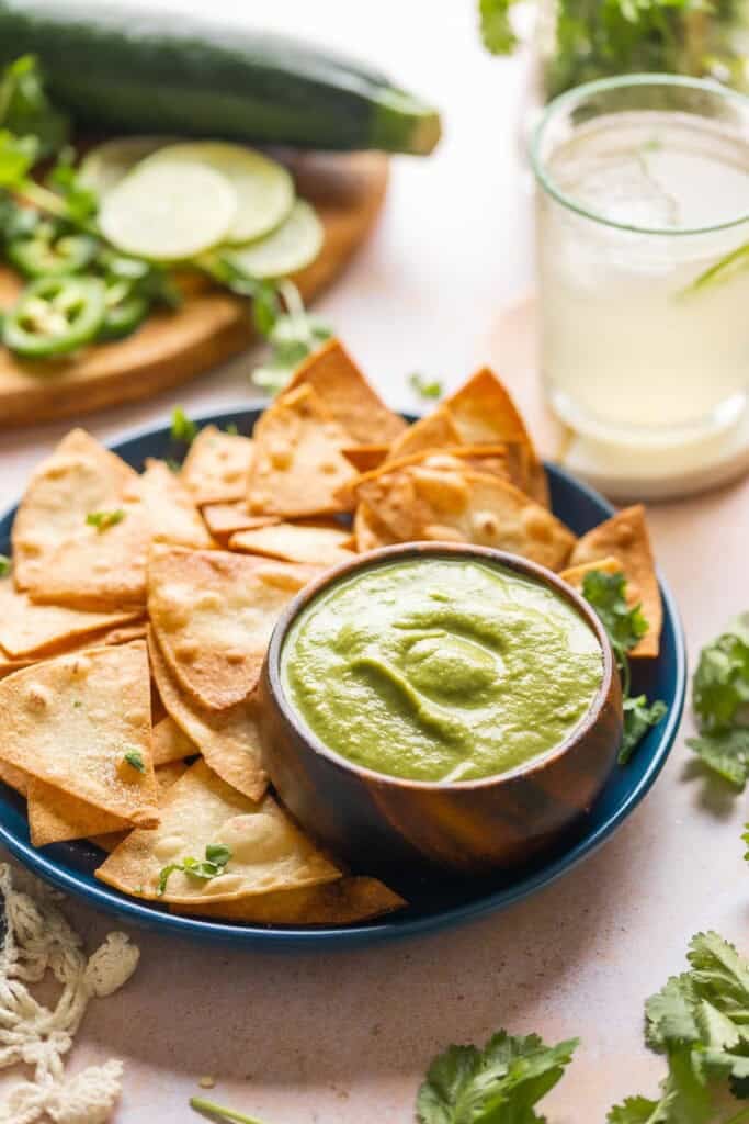 Jalapeño Zucchini Salsa in a dip bowl on a plate with tortilla chips with fresh cilantro, zucchini and Jalapeño on a cutting board in the back and margarita on the side.