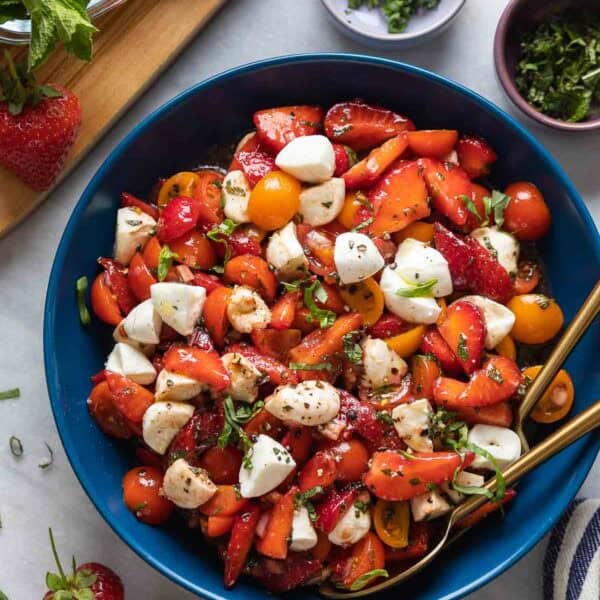 Strawberry Tomato Balsamic Salad in a serving bowl with fresh ingredients surrounding the dish. Salad is made with sliced strawberries, cherry tomatoes, mozzarella and fresh mint and basil.
