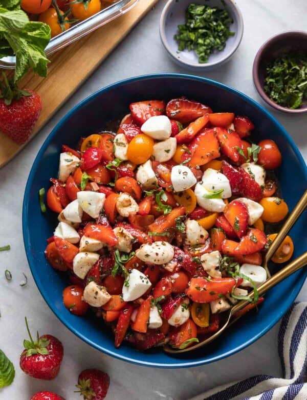 Strawberry Tomato Balsamic Salad in a serving bowl with fresh ingredients surrounding the dish. Salad is made with sliced strawberries, cherry tomatoes, mozzarella and fresh mint and basil.