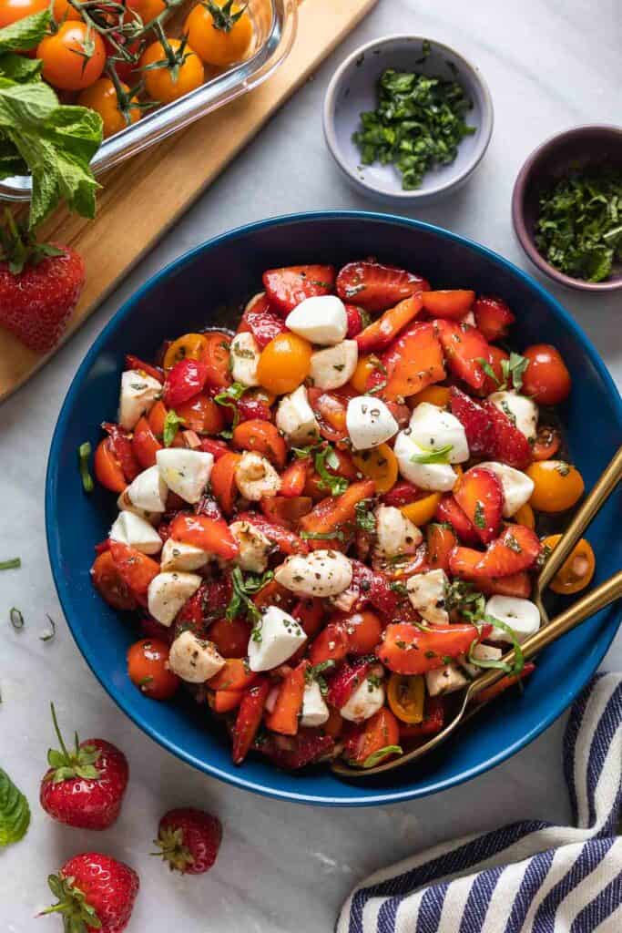 Strawberry Tomato Balsamic Salad in a serving bowl with fresh ingredients surrounding the dish. Salad is made with sliced strawberries, cherry tomatoes, mozzarella and fresh mint and basil.