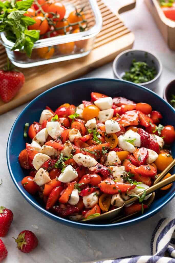 A bowl full of Strawberry Tomato Balsamic Salad with serving spoons and the fresh ingredients around the bowl.