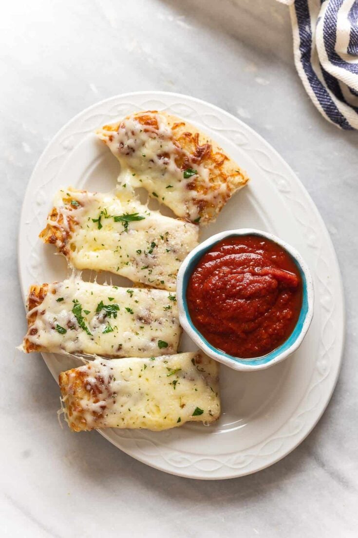 Four slices of Gluten Free Cheesy Breadsticks and a small bowl of marinara sauce on a plate.