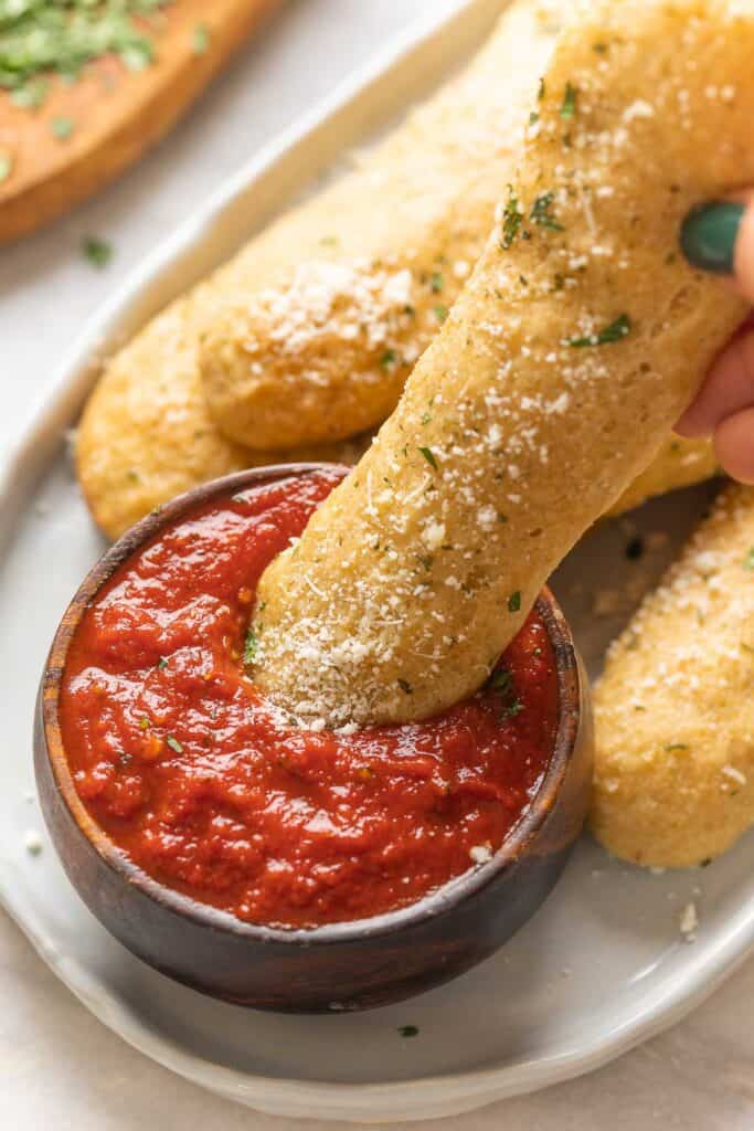A gluten free breadstick dipping into a small bowl of marinara sauce.