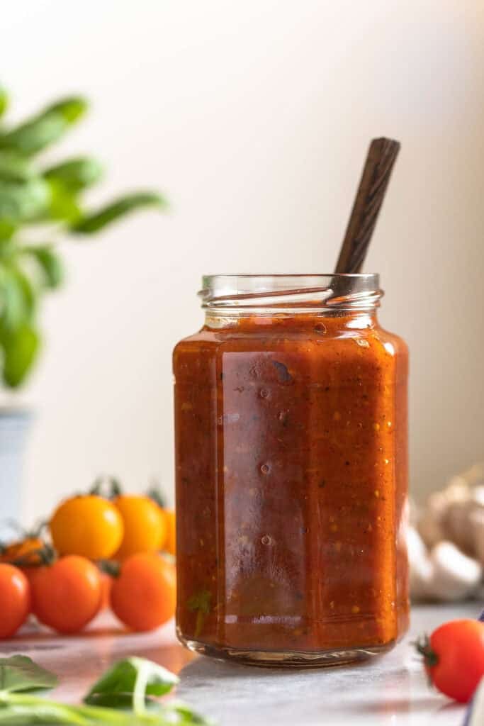 A jar full of homemade Homemade Pizza Sauce with cherry tomatoes, fresh basil and garlic in the back.