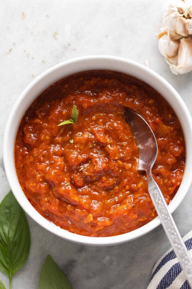 Homemade Pizza Sauce in a bowl with a couple fresh basil leaves and a spoon. Garlic head and fresh basil spring on the side.