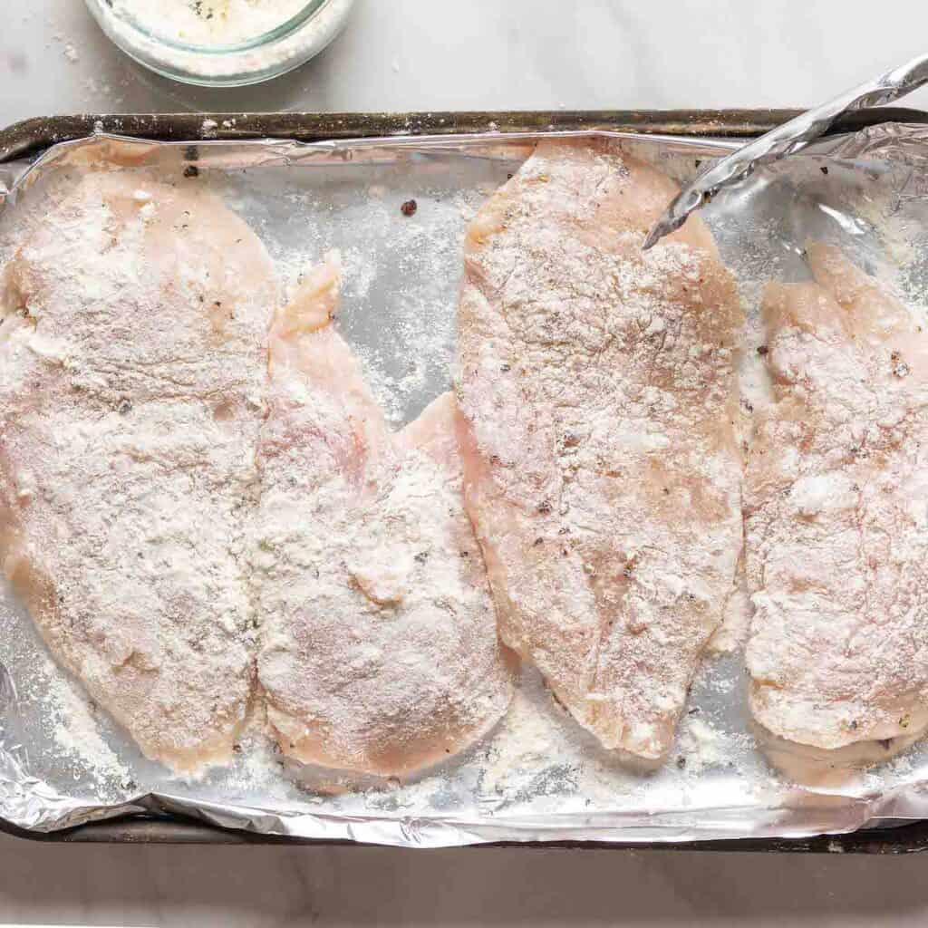 Four, raw chicken breast coated in salt, pepper and gluten free flour on a foil-lined baking sheet.