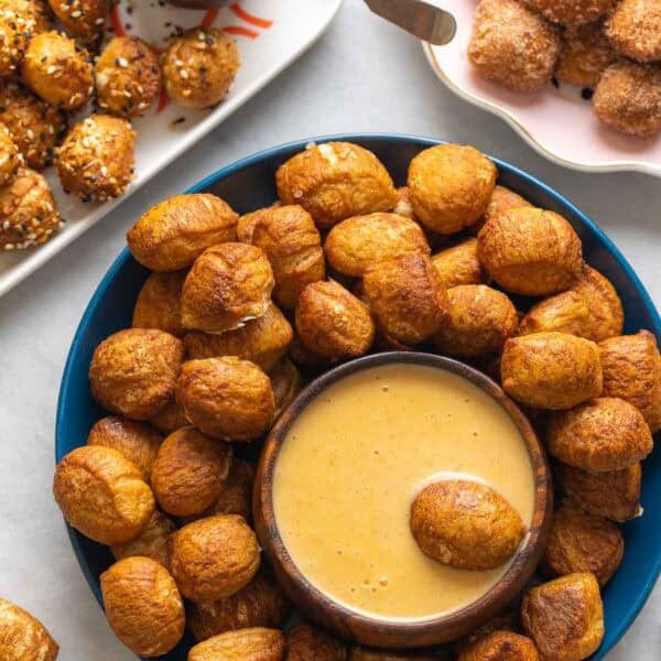Gluten Free Pretzel Bites on a plate with a bowl of cheese sauce and aa pretzel bite in the cheese sauce. Pretzel bites topped with everything bagel seasoning on one plate with a bowl of grain mustard in one corner and a plate of cinnamon sugar coated pretzel bites in the other corner.