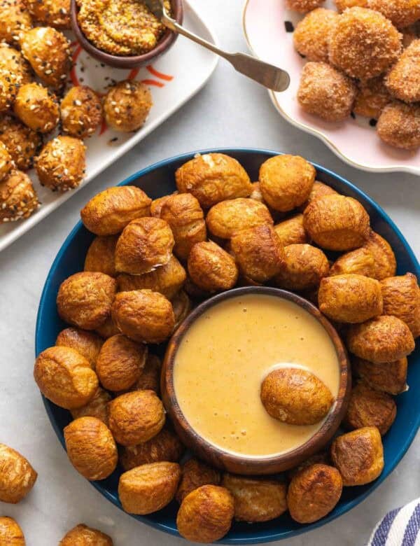 Gluten Free Pretzel Bites on a plate with a bowl of cheese sauce and aa pretzel bite in the cheese sauce. Pretzel bites topped with everything bagel seasoning on one plate with a bowl of grain mustard in one corner and a plate of cinnamon sugar coated pretzel bites in the other corner.
