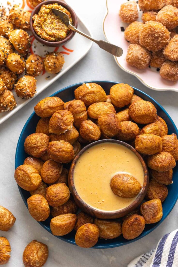 Gluten Free Pretzel Bites on a plate with a bowl of cheese sauce and aa pretzel bite in the cheese sauce. Pretzel bites topped with everything bagel seasoning on one plate with a bowl of grain mustard in one corner and a plate of cinnamon sugar coated pretzel bites in the other corner.