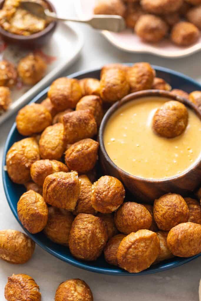 Gluten Free Pretzel Bites on a plate with a bowl of cheese sauce and aa pretzel bite in the cheese sauce. A small bowl of grain mustard and another plate of cinnamon sugar pretzel bites in the back.
