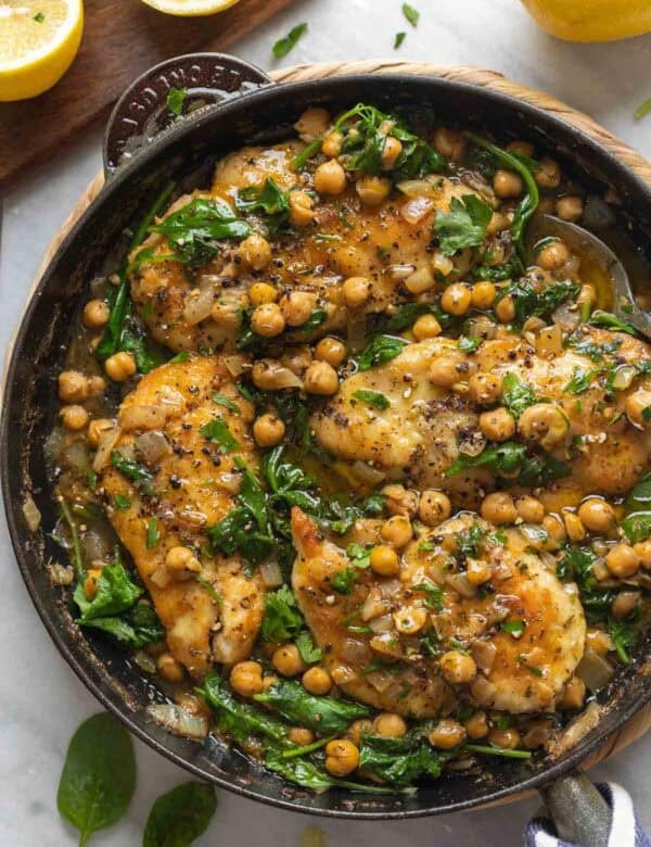 Pan seared chicken breast mixed into a skillet full of lemon-anchovy simmer chickpeas and wilted spinach with a spoon inside the pan.