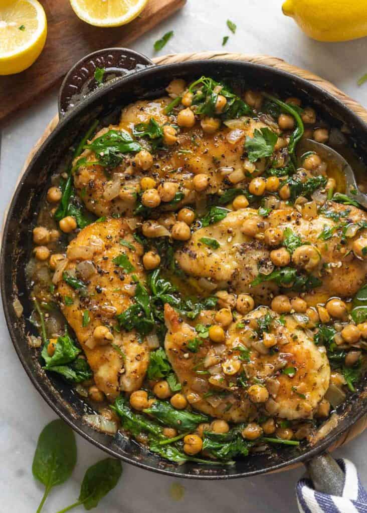 Pan seared chicken breast mixed into a skillet full of lemon-anchovy simmer chickpeas and wilted spinach with a spoon inside the pan.