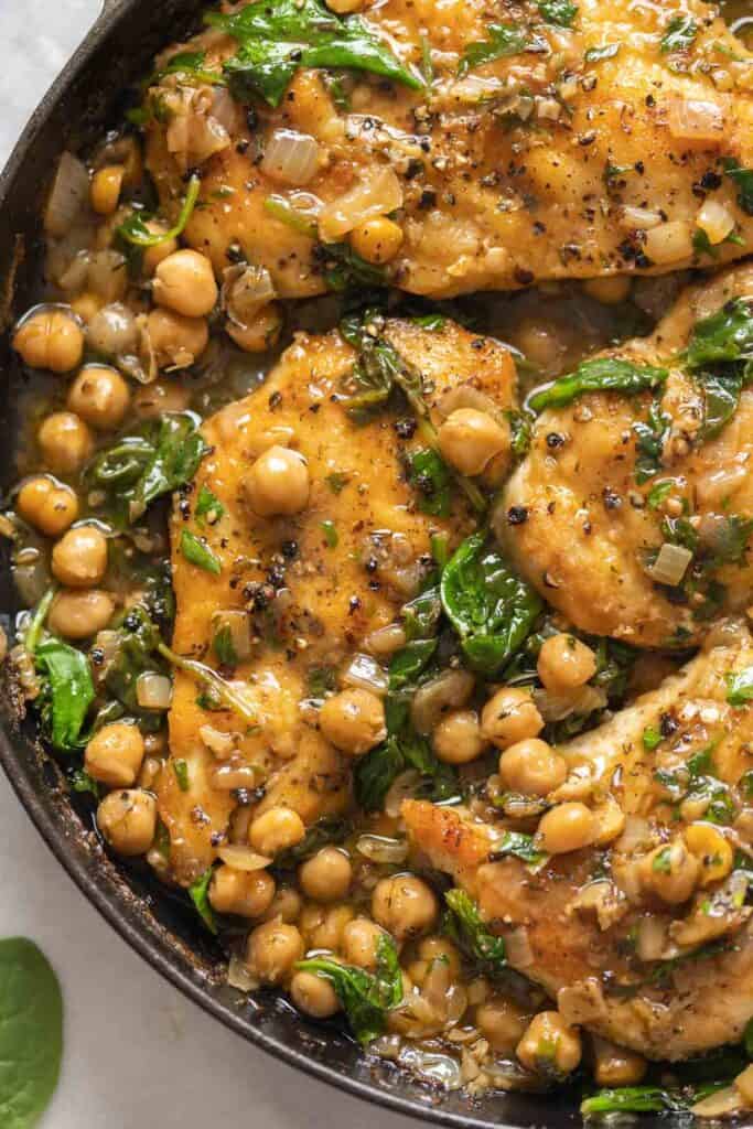 Pan seared chicken breast nestled inside a cast iron skillet full of chickpeas simmered in a lemon-anchovy sauce with cracked pepper on top.