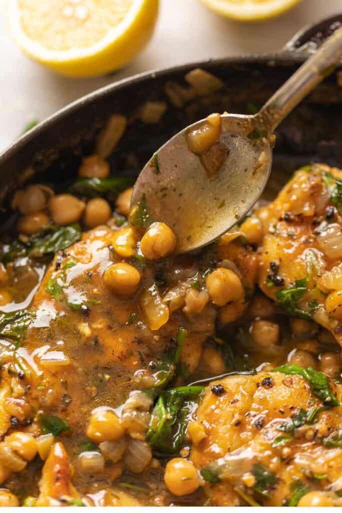 A spoon pouring lemon-anchovy chickpeas over pan seared chicken breast in a skillet.