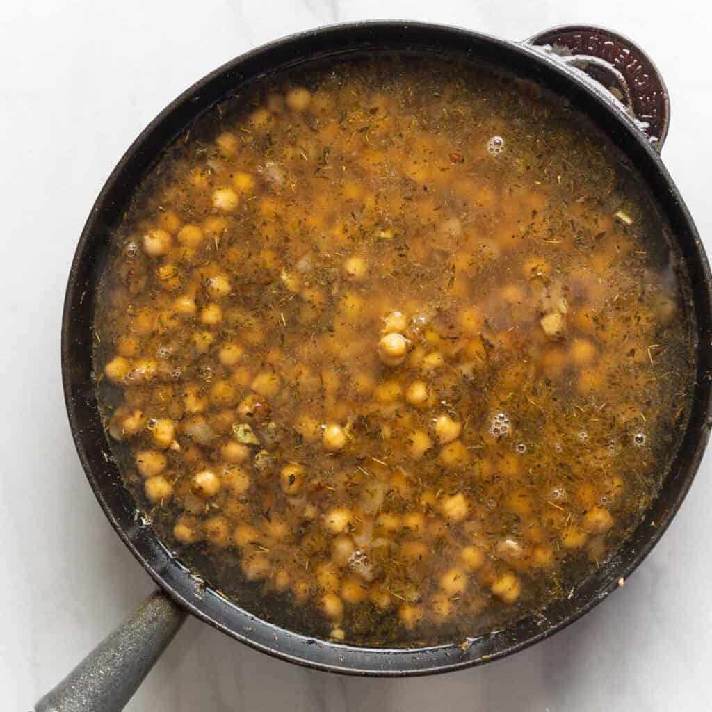 Chicken stock and lemon juice covering sautéed chickpeas and aromatics inside a skillet.