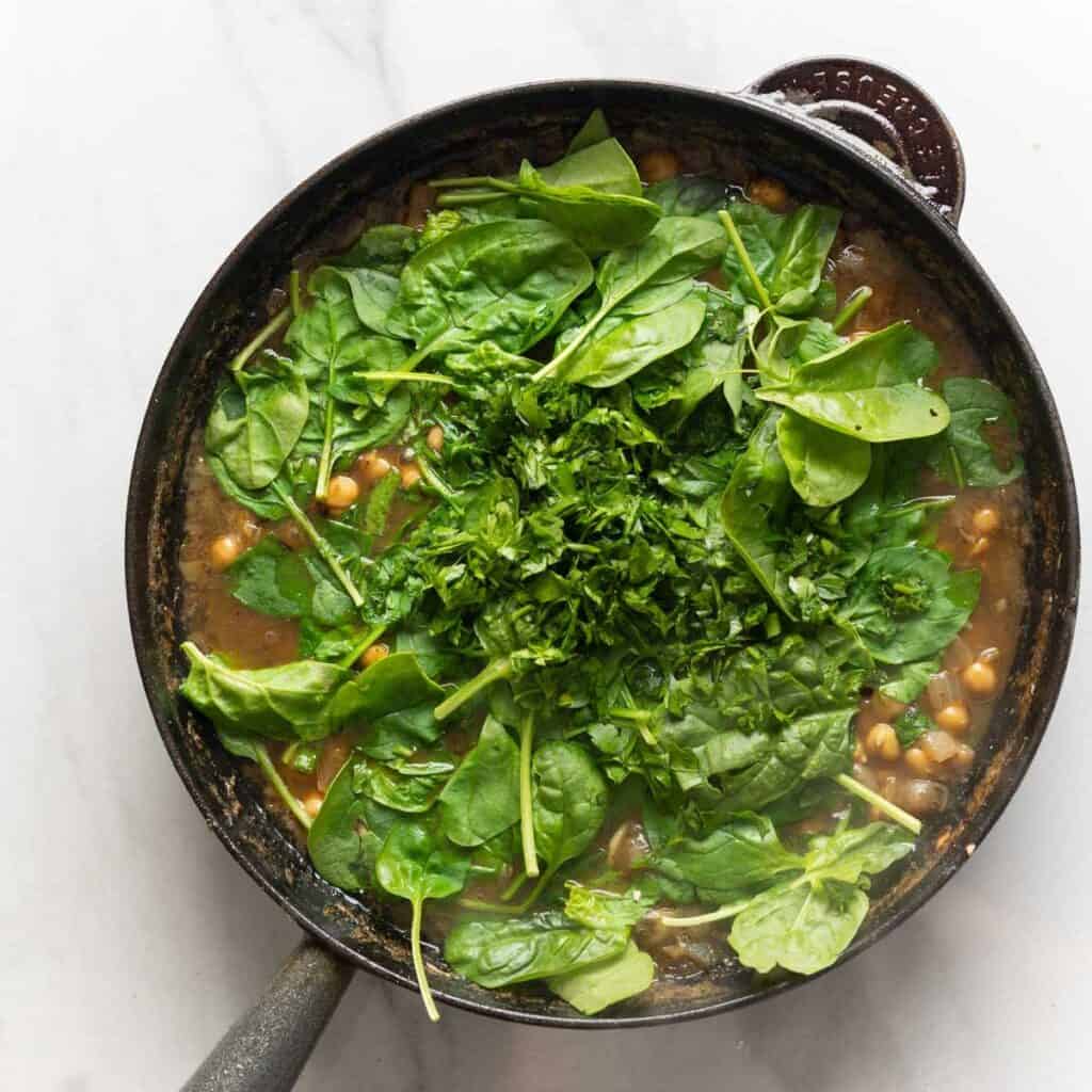 Fresh spinach and parsley on top of a skillet full of a lemon-anchovy sauce with chickpeas.
