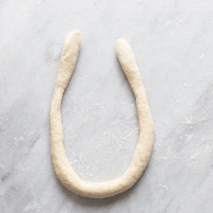 A gif showing how to shape pretzel dough: make a U-shape, twist the top ends around each other twice, pull the end down toward you, straighten out the ends and loops.