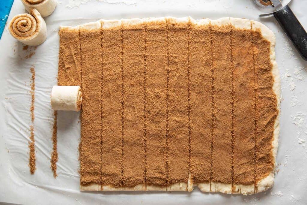 Gluten free cinnamon roll dough rolled out in a big rectangle then covered with a butter, cinnamon-sugar mixture and cut into strips to roll up into cinnamon rolls.
