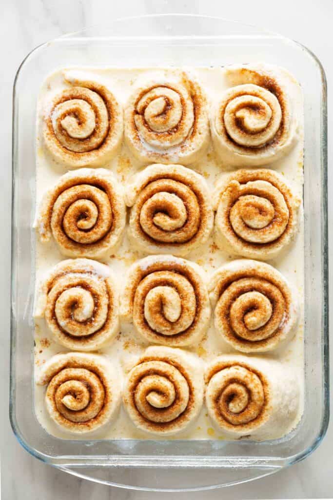 A glass baking dished filled with 12 unbaked gluten free cinnamon rolls after they've had time to proof & puff up. Heavy cream poured into the dish to bake with the cinnamon rolls.