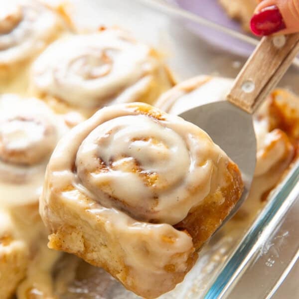 A gluten free cinnamon roll covered in a cream cheese frosting held up over a glass baking dish on a metal spatula.