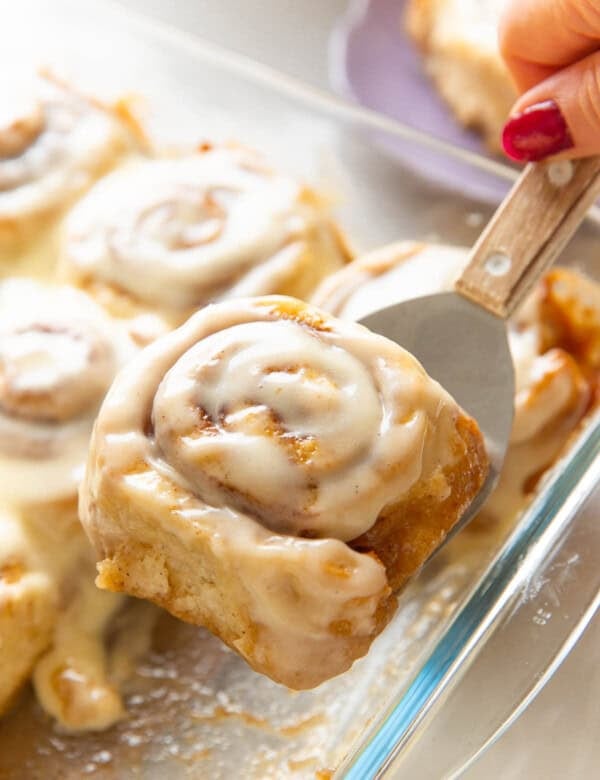 A gluten free cinnamon roll covered in a cream cheese frosting held up over a glass baking dish on a metal spatula.