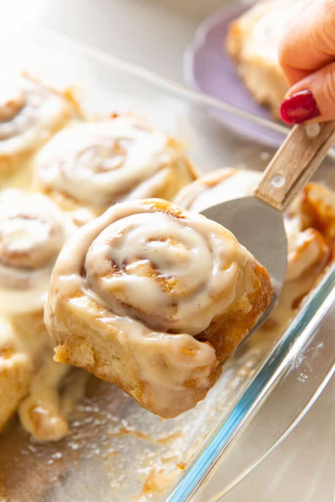 A gluten free cinnamon roll covered in a cream cheese frosting held up over a glass baking dish on a metal spatula.