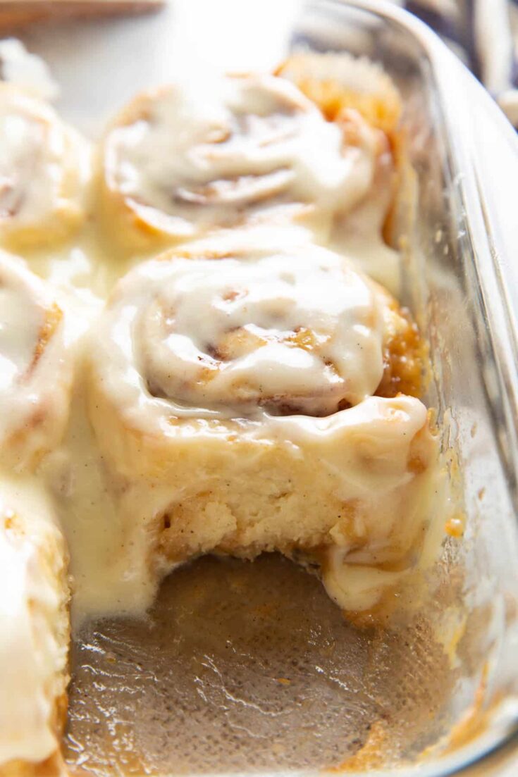 Gluten free cinnamon rolls covered in a cream cheese frosting in a glass baking dish. One cinnamon rolls removed from the dish to provide a side on view of another cinnamon roll.
