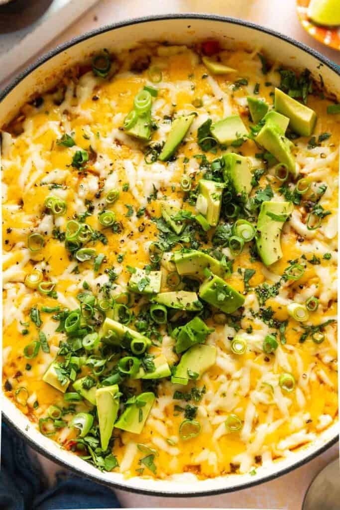 A casserole dish full of cheesy chicken enchilada rice with green onions, fresh cilantro and sliced avocado on top. 