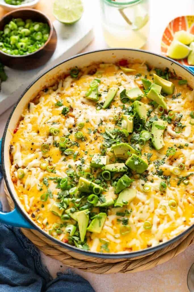 A casserole dish full of cheesy chicken enchilada rice with green onions, fresh cilantro and sliced avocado on top. Lime wedges and a bowl of spring onions in the back.