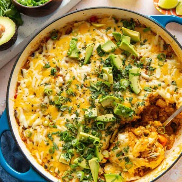 A casserole dish full of cheesy chicken enchilada rice with a serving spoon. Casserole garnished with green onions, fresh cilantro and sliced avocado on top.
