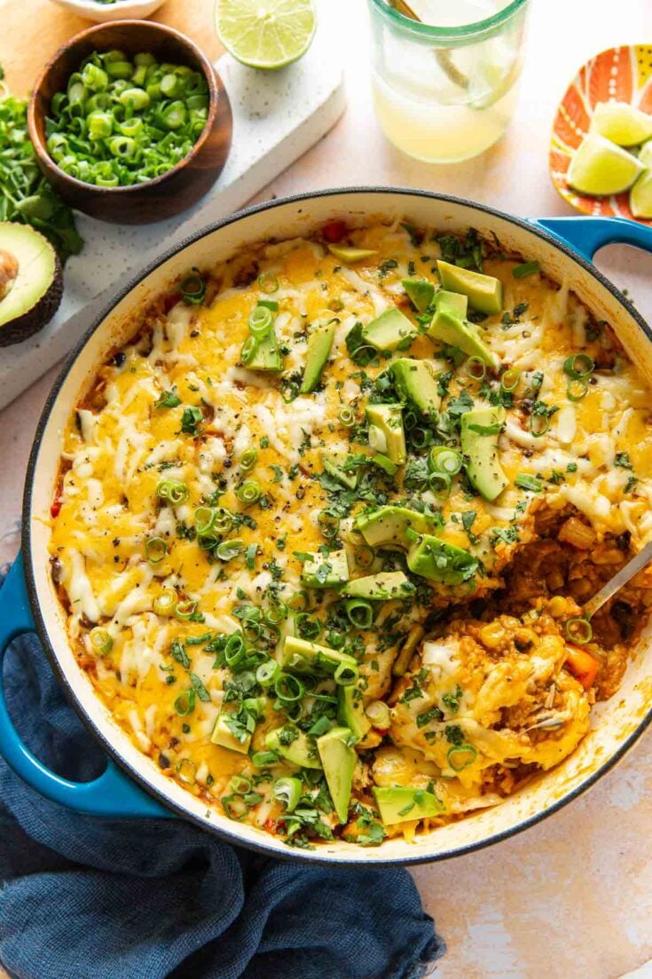 A casserole dish full of cheesy chicken enchilada rice with a serving spoon. Casserole garnished with green onions, fresh cilantro and sliced avocado on top.