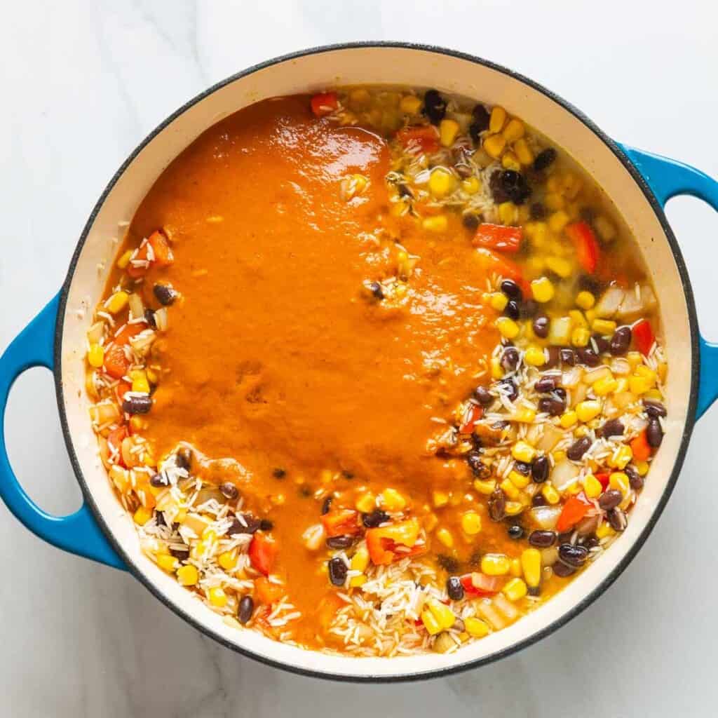 Red enchilada sauce and chicken stock added to a casserole dish full of mixed rice, beans, corn and veggies.