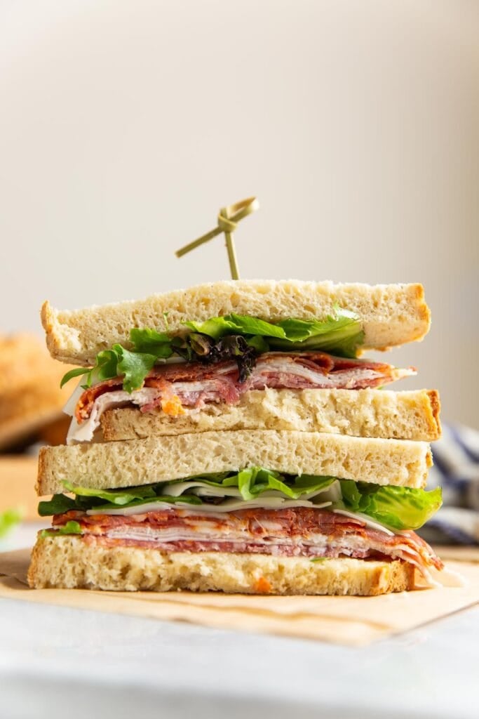 A sandwich made with gluten free bread filled with cheese, deli meats and salad leaves cut in half and stack on top of each other.