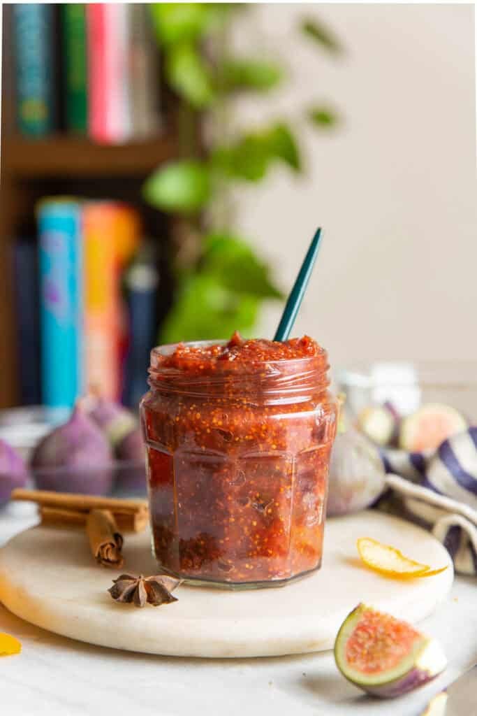 A small jar full of homemade fig jam surrounded by fresh figs, dried orange peel, star anise and cinnamon sticks.