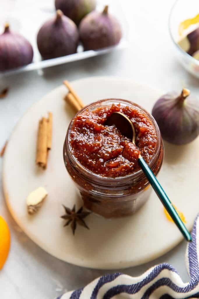 A small jar full of homemade fig jam surrounded by fresh figs, dried orange peel, a piece of ginger, star anise and cinnamon sticks and a small spoon in the jar.