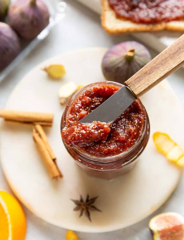 A small butter knife placed over a jar of homemade fig jam with the fresh ingredients surrounding the jar.
