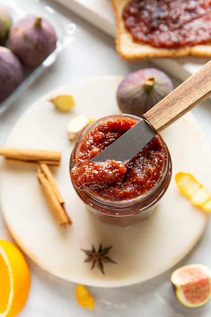 A small butter knife placed over a jar of homemade fig jam with the fresh ingredients surrounding the jar.