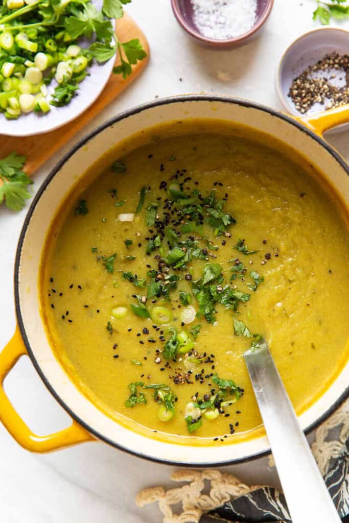 A dutch oven full of a pureed aloo gobi soup (curried potato and cauliflower soup) with a soup ladle in the pot. The soup is topped with sliced spring onions, chopped cilantro, black pepper and nigella seeds.