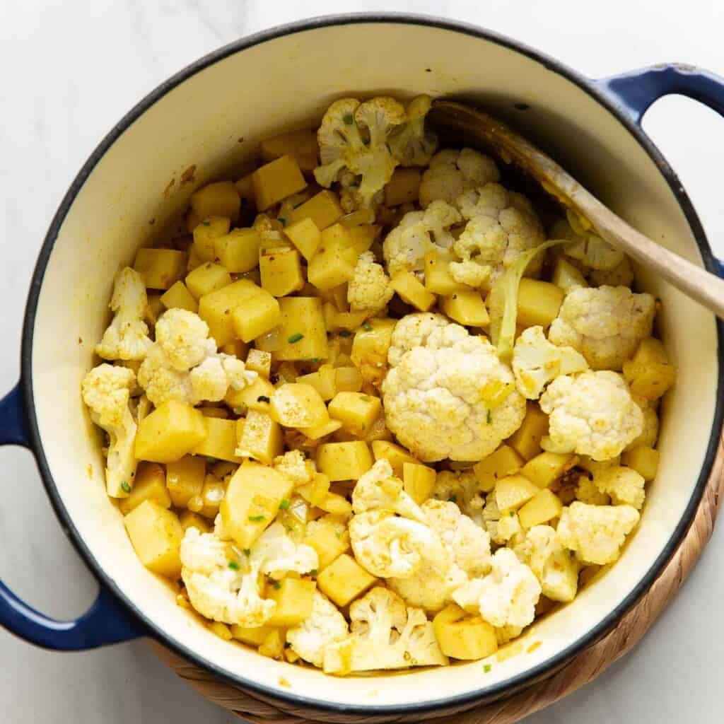 A large dutch oven pot full of diced potato and cauliflower florets mixed into a spiced onion mixture.