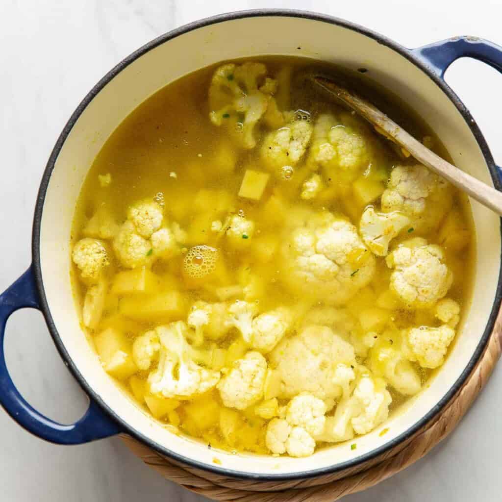 A large dutch oven pot full of vegetable broth covering diced potato and cauliflower florets mixed into a spiced onion mixture.