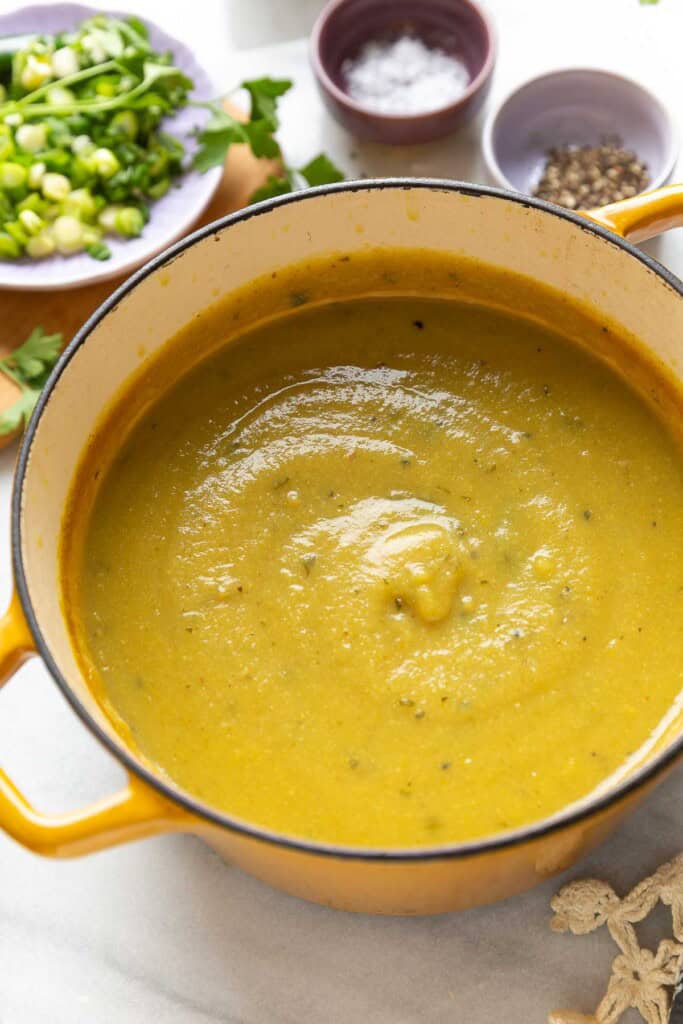 A dutch oven full of a pureed aloo gobi soup (curried potato and cauliflower soup) with fresh garnished, salt and pepper in the back.