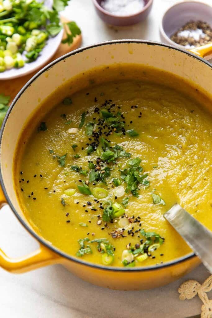 A dutch oven full of a pureed aloo gobi soup (curried potato and cauliflower soup) topped with sliced spring onions, chopped cilantro, black pepper and nigella seeds.