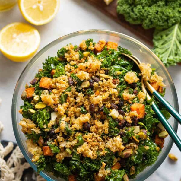 A salad made with roasted sweet potato & black beans, kale, cheese and chopped pistachios lot crispy parmesan quinoa in a glass salad bowl with serving spoons surrounded by the raw ingredients.
