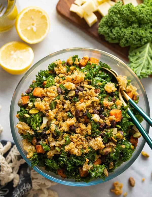 A salad made with roasted sweet potato & black beans, kale, cheese and chopped pistachios lot crispy parmesan quinoa in a glass salad bowl with serving spoons surrounded by the raw ingredients.